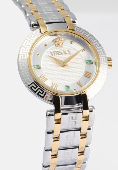 Orologio Versace con bracciale in acciaio inossidabile oro e argento, quadrante rotondo bianco, dettagli verdi, logo del leone in rilievo e motivo geometrico sulla lunetta.