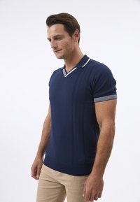Marineblaues Strickpolo mit V-Ausschnitt, mit weißen Streifenakzenten am Kragen und an den Ärmeln sowie strukturierten vertikalen Linien auf der Vorderseite.