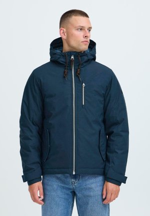 Man wearing a navy blue hooded jacket with front zipper and chest pocket, paired with blue jeans, looking to the side against white background.