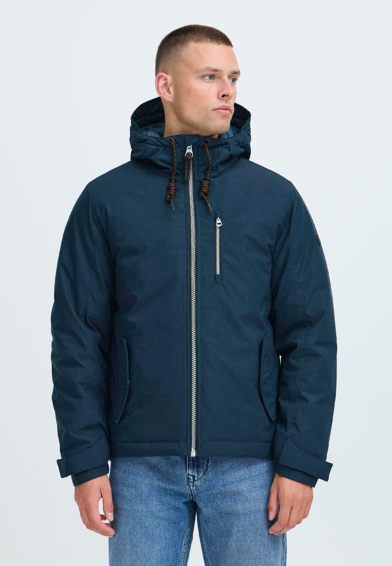 Uomo che indossa una giacca con cappuccio blu navy con zipper frontale e tasca sul petto, abbinata a jeans blu, che guarda di lato su sfondo bianco.