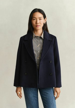 Femme aux cheveux raides et foncés portant un manteau croisé bleu marine foncé, une chemise à carreaux boutonnée et un jean bleu sur un fond uni.