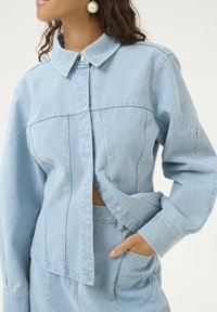 Giacca di denim azzurro chiaro con colletto strutturato e chiusura a bottone a pressione. Presenta tasche laterali ed è leggermente accorciata in vita.