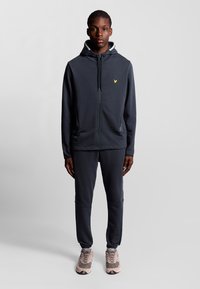 Donkergrijze zip-up hoodie met een groen logo op de borst, gecombineerd met bijpassende joggers en lichtgekleurde sportschoenen. Glad textuur overal.