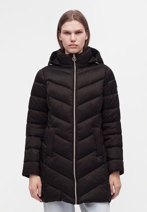 Femme portant une longue doudoune noire matelassée avec capuche et fermeture éclair à l'avant, debout devant un fond clair uni.