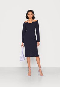 Vestido azul marinho de manga longa com design off-shoulder, silhueta ajustada e comprimento médio. Combinado com uma mala lavanda e sapatos de salto combinando.