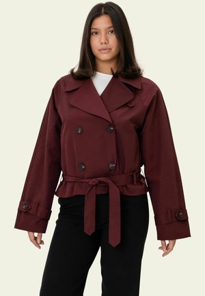 Noor naine, kes kannab burgundia värvi topeltkaravanist jopet vööga vöökohal, musti pükse ja valget sätendust ühevärvilise heleda tausta ees.