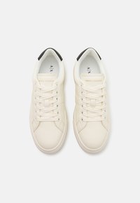 Kremfargede lær-sneakers med hvite snørebånd og en teksturert overflate. Svart detaljer på hælen og merkevaren inne i skoen.