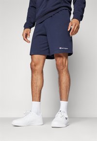 Pantalones cortos de algodón azul marino con cintura elástica, que presentan el logo de "Champion" en la pierna izquierda. Combinados con zapatillas blancas y calcetines.