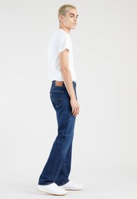 Jauns vīrietis ar īsiem gaišiem matiem, tērpies baltā T-kreklā, tumši zilās džinsās un baltos sporta apavos, stāv profila pozā uz vienkrāsaina fona.