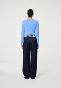 Vrouw gezien van achteren in een lichtblauwe fluffy trui, met een wijde spijkerbroek, een koeienprintriem en een schelpenketting, met haar in een lage bun.