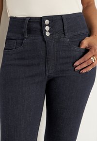 Höga mörk denimjeans med tre silverknappar, framfickor och en slät textur. Har en figurnära design.