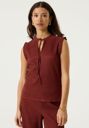Vrouw staand, draagt een mouwloos, geribd bordeauxrood topje met strikdetail bij de hals en bijpassende broek, gouden oorringen en een armband.