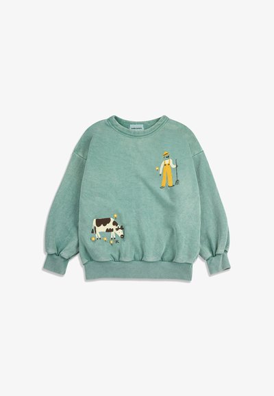 Sweatshirt vert avec des illustrations d'un fermier tenant une fourche et d'une vache noire et blanche en train de paître, accompagnée de petites fleurs jaunes.