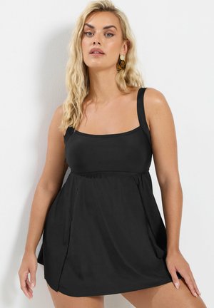 Blonde vrouw die een zwarte mouwloze jurk met een vierkante halslijn en grote schildpad- en gouden oorbellen draagt, staand tegen een witte muur.