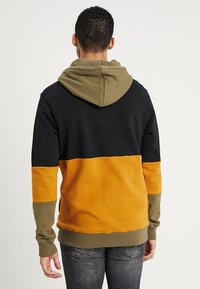 Kleurgeblokkeerde hoodie met een zwarte bovenkant, oranje midden en olijfgroene accenten. Zacht materiaal met een buidelzak en een verstelbare capuchon.