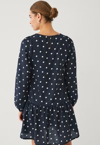 Vestido azul marino con lunares, mangas largas abullonadas, falda en capas y cierre de botones en el cuello. Fabricado con una tela ligera y texturizada.