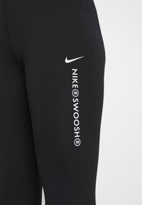 Svart atletisk långärmad tröja med en tight passform. Visar en vit Nike-logotyp och texten "NIKE®SWOOSH®" på ärmen. Slät tygtextur.