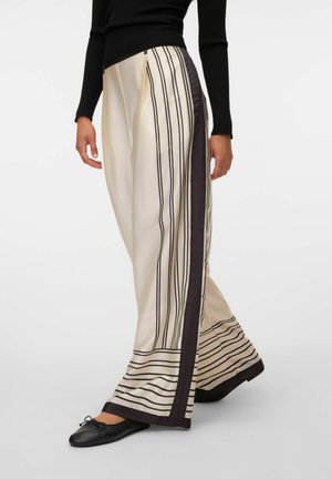 Femme portant un pantalon large crème avec des rayures noires verticales et horizontales et des ballerines noires, associée à un haut noir à manches longues.