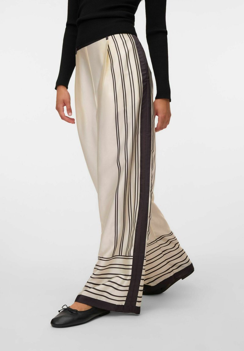 Femme portant un pantalon large crème avec des rayures noires verticales et horizontales et des ballerines noires, associée à un haut noir à manches longues.
