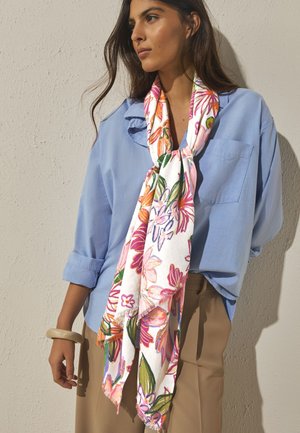 Frau trägt hellblaues Hemd und beige Hose mit einem weißen Blumenschal um den Hals gebunden und einem beigen Armreif am Handgelenk.