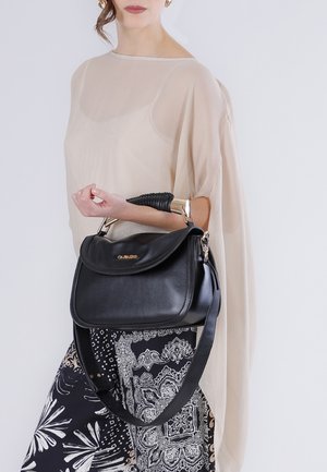 Donna che indossa un top beige trasparente e pantaloni a motivo bianco e nero, che tiene una borsa di pelle nera con dettagli dorati.