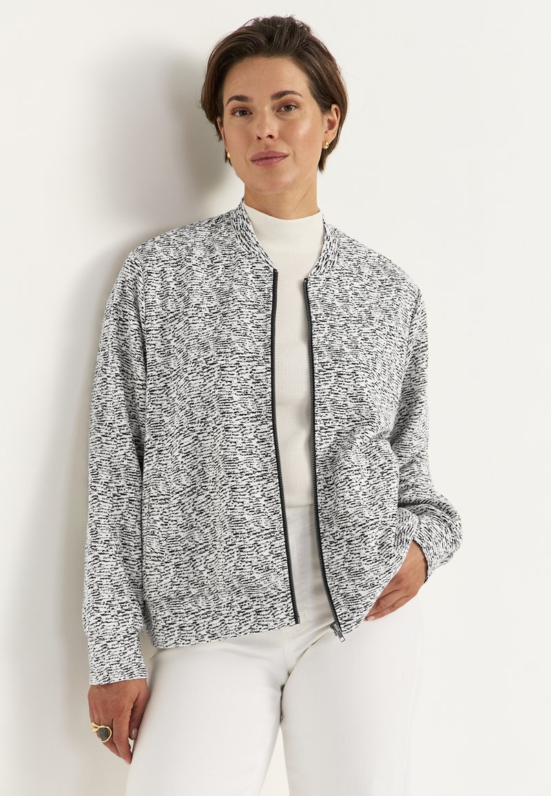 Sort-hvid mønstret bomberjakke med lynlåslukning, ribbet krave og løs pasform, båret over en hvid højhalset top.