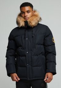 Schwarze Pufferjacke mit einem mit Fell gefütterten Kapuze. Verfügt über zwei Fronttaschen, Druckknöpfe und ein gesticktes Logo am Ärmel. Weiches, isoliertes Material.