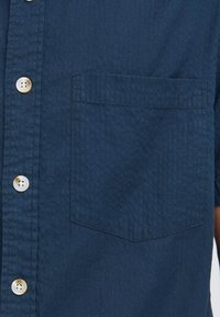 Camisa de botón de manga corta en tela azul oscura, con un bolsillo con solapa y cierre de botones de color claro. Superficie texturizada con patrones sutiles.