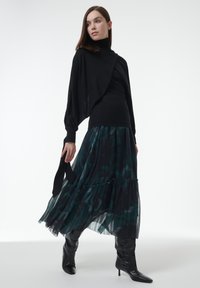Zwarte gebreide trui met gedrapeerd ontwerp, gecombineerd met een vloeiende teal en zwarte geplooide rok. Model draagt kniehoog zwarte haklaarzen.
