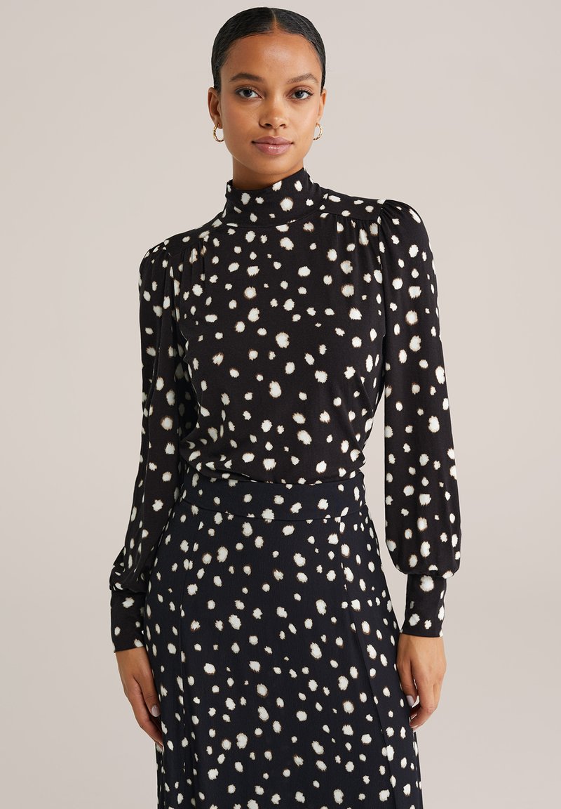 Kobieta z zaczesanymi do tyłu włosami, nosząca złote kolczyki koła oraz czarną sukienkę z długim rękawem w białe nieregularne kropki na neutralnym tle.