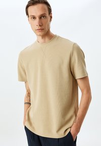 Beige T-Shirt mit kurzen Ärmeln aus strukturiertem Material und Rundhalsausschnitt, mit einem dezenten Nahtdetail auf der Vorderseite und lockerer Passform.