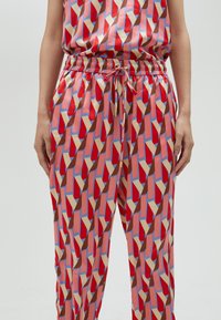 Red and pink patterned pants with geometric shapes in blue, beige, and brown; elastic waistband with drawstring; soft fabric texture.