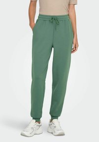 Zelene sweatpants od mekog materijala, s elastičnim pojasom s utezima, bočnim džepovima i sužavajućim manžetama na gležnjevima.