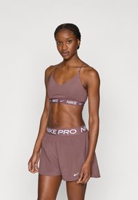 Αθλητικό σουτιέν σε σκούρο μωβ χρώμα με λεπτές τιράντες και λωρίδα με λογότυπο· αντίστοιχα σορτς σε παρόμοια απόχρωση, με ελαστική μέση που φέρει το κείμενο "NIKE PRO".