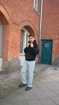 Man in een zwarte hoodie en lichtblauwe spijkerbroek die zijn kraag aanpast, staand op een kinderkopjespad naast een rood bakstenen gebouw met een boogvormig raam.