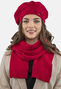 Red knitted beret with floral accents and a matching scarf, both featuring a smooth texture and ribbed edges, worn over a beige coat.