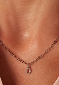 Zilveren ketting met verlengde schakels en een kleine, hoofdletter "D" hanger, rustend tegen gebruinde huid.