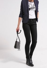 Merimiespuku sininen bleiseri valkoisen graafisen t-paidan päällä; mustat kapealahkeiset farkut; mustat nilkkurit; kädessä pieni harmaa laukku; vähäiset asusteet.