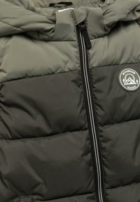 Chaqueta acolchada de color verde oliva oscuro, con patrón acolchado vertical, cremallera negra con dos detalles plateados y parche de logo circular.