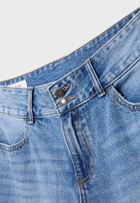 Niebieska sekcja talii dżinsów wykonanych z denimu, pokazująca podwójne metalowe guziki, szlufki na pasek oraz przednie kieszenie z pomarańczowym przeszyciem.