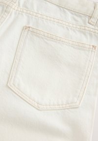 Nahaufnahme eines cremefarbenen Denim-Stoffs, der eine Gesäßtasche mit beigen Nähten und eine sichtbare Gürtelschlaufe oben rechts zeigt.