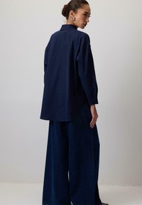 Camicia blu navy con bottoni e maniche lunghe, dettaglio della plissettatura sul retro e orlo arrotondato abbinata a pantaloni in denim scuro a gamba larga.