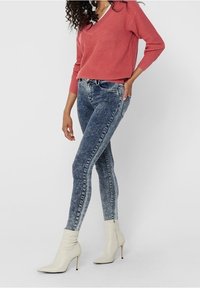 Roodroze gebreide trui met V-hals, gecombineerd met blauwe skinny jeans van medium was, aangevuld met witte stiletto enkellaarsjes.