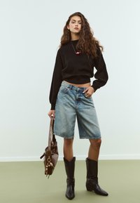 Žena s dugom kovrčavom kosom, u crnom puloveru, trapericama do koljena, s crnim kaubojskim čizmama, drži smeđu torbicu s privjeskom.
