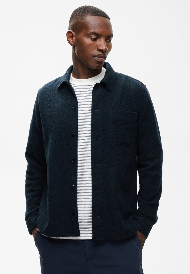 Homme portant une veste bleu marine foncé à boutons sur une chemise blanche et grise rayée, regardant vers le bas et sur le côté avec les mains dans les poches.