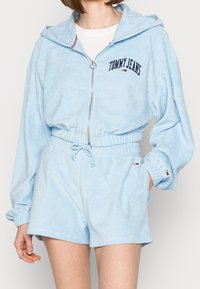 Light blue velour hoodie with a zip and drawstring waist, featuring "TOMMY JEANS" logo. Elastic cuffs and shorts with minimal detailing.