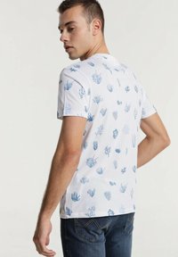 Weißes T-Shirt mit Mustern aus blauen Korallen und Seegras. Kurze Ärmel, Rundhals und lockere Passform. Trägt man mit dunkelblauen Jeans.