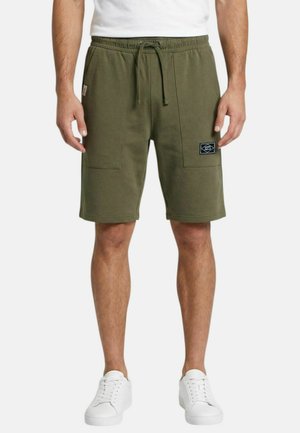 Homme portant un short kaki arrivant aux genoux avec poches à l'avant, une taille à cordon, des baskets blanches et une chemise blanche.