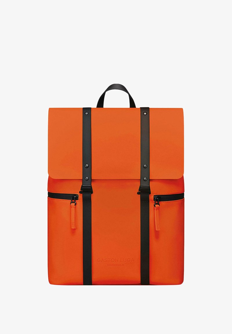 Oranžs mugursoma ar salokāmu vāku, melniem siksnām un rāvējslēdzējiem. Izgatavota no gludas materiāla, ar priekšējo kabatu un logotipa detaļām.