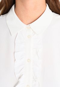 Blusa bianca con colletto a punta, caratterizzata da una placca frontale arricciata e bottoni simili a perle; tessuto liscio con una consistenza morbida.